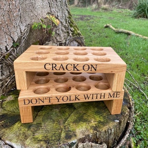 Puede incluir: Un soporte para huevos de madera de dos niveles. El nivel superior tiene el texto "CRACK ON" y el nivel inferior tiene el texto "DON'T YOLK WITH ME".