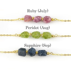 May include: Three gold chains with different birthstone beads. The top chain has three pink ruby beads, the middle chain has four green peridot beads, and the bottom chain has four blue sapphire beads. The chains are labeled with the birthstone name and month.