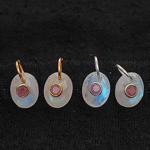 May include: Four oval-shaped earrings with a central pink gemstone. Two earrings have gold-colored hoops, and two have silver-colored hoops. The earrings have a pearlescent, iridescent appearance, set against a black background.