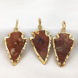 May include: Three arrowhead pendants with gold-coloured trim. The pendants have a reddish-brown stone centre with white and tan inclusions. Each pendant has a gold-coloured loop at the top for hanging.