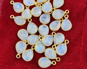 May include: A group of 18 teardrop-shaped moonstone gemstone pendants with gold-plated jump rings. The stones are a milky white with a slight iridescent shimmer.