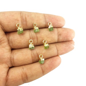 May include: Several small, raw, green gemstone charms with gold-coloured metal loops. The gemstones have a rough, natural texture and are irregularly shaped. The charms are displayed against a light background.