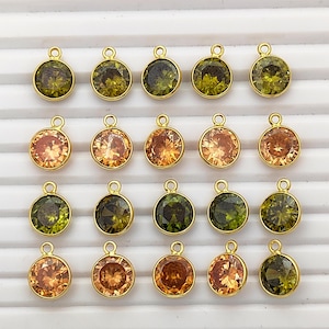 Könnte beinhalten: Eine Sammlung runder, goldfarbener Anhänger mit farbigen, facettierten Edelsteinen. Die Edelsteine sind in Grüntönen, Orange und Bernstein gehalten. Jeder Anhänger hat oben eine kleine Schlaufe zum Befestigen an einer Kette oder einem anderen Schmuckbestandteil.