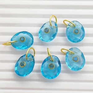 Pode incluir: Seis pingentes facetados, translúcidos, de cor azul brilhante e formato oval, cada um com um pequeno detalhe circular com borda dourada no centro. Cada pingente inclui uma argola de metal dourado no topo para fixação.