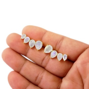 Może przedstawiać: Para złotych kolczyków z motywem łzy księżycowego kamienia. Każdy kolczyk ma pięć księżycowych kamieni w stopniowanych rozmiarach, z największym kamieniem na dole.