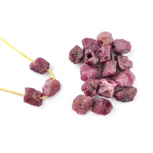 May include: A pile of rough, pink ruby gemstones, some with drilled holes, and a gold chain with four ruby beads.
