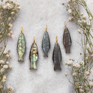 May include: Five carved fish pendants with gold-colored loops for hanging. The pendants are in shades of gray, green, and blue, with detailed scale patterns. The pendants are displayed on a white surface with sprigs of white flowers.