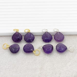 May include: A collection of amethyst gemstone pendants. Each pendant is carved in a shell shape with detailed ridges. The pendants have gold and silver-colored loops for attaching to a chain or cord. The amethyst color is a deep, rich purple.