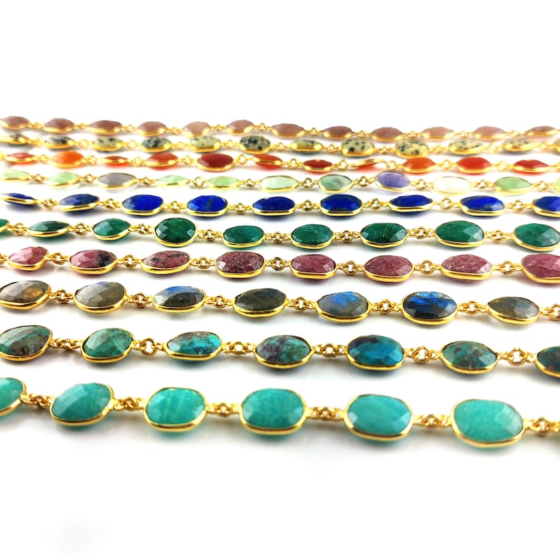 May include: A close-up of a gold chain with oval-shaped gemstones in various colors, including red, green, blue, and turquoise. The chain is laid out in rows, showcasing the different gemstone colors.