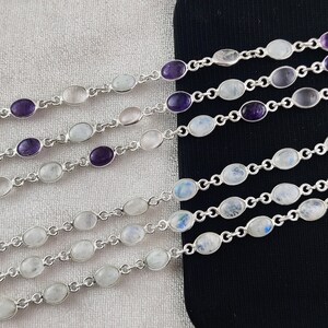 May include: Five silver bracelets with oval-shaped moonstone and amethyst gemstones. The bracelets are layered on a black surface.