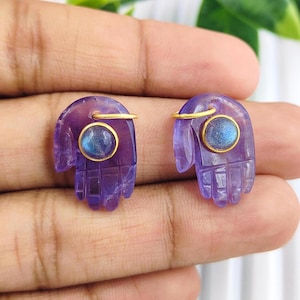 Puede incluir: Un par de pendientes de mano de hamsa de amatista con detalles dorados y una piedra preciosa redonda de labradorita en el centro de cada mano.