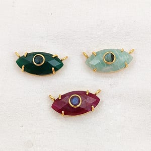 May include: Three gemstone eye pendants with gold-colored metal accents. The gemstones are emerald green, light green, and ruby red, each with a contrasting center stone. The pendants are designed with a faceted cut and are suitable for jewelry making.