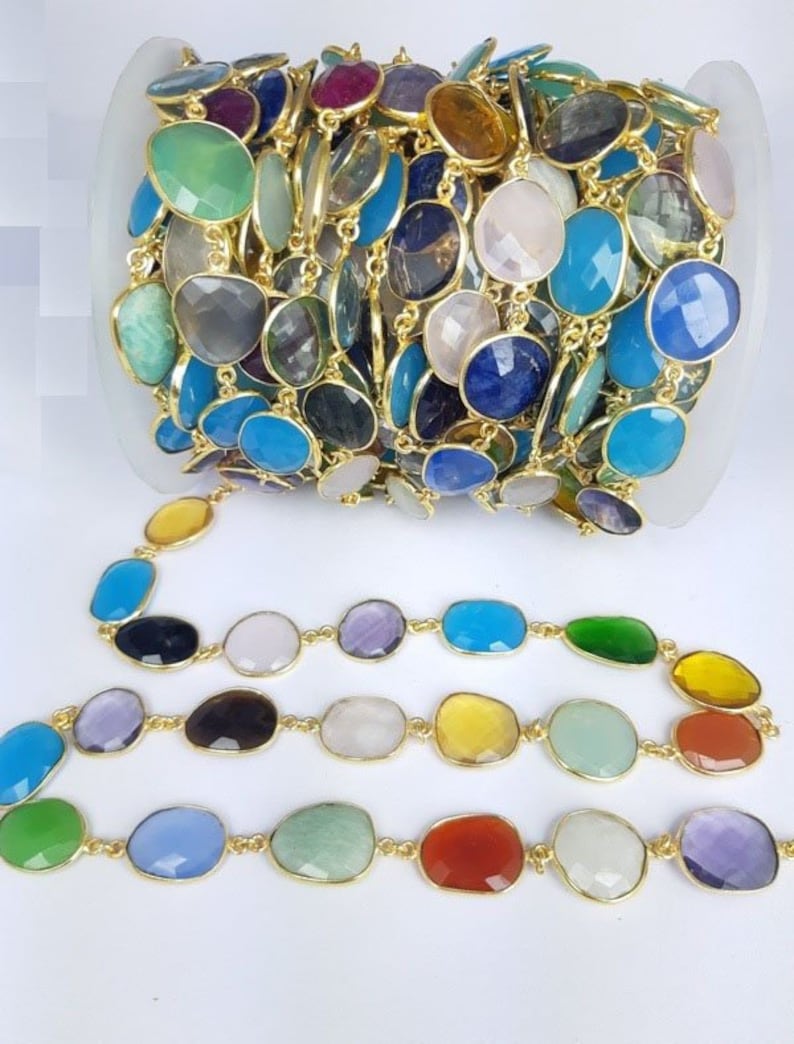 May include: A roll of colorful gemstone chain with gold-colored links. The gemstones are oval-shaped and come in various colors, including blue, green, purple, and yellow. The chain is displayed on a white surface.