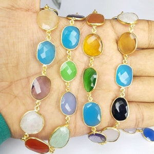 May include: A multi-colored gemstone necklace with gold-colored metal links. The necklace features a variety of faceted, oval-shaped gemstones in shades of blue, green, yellow, and purple. The gemstones are approximately 1 inch in length.
