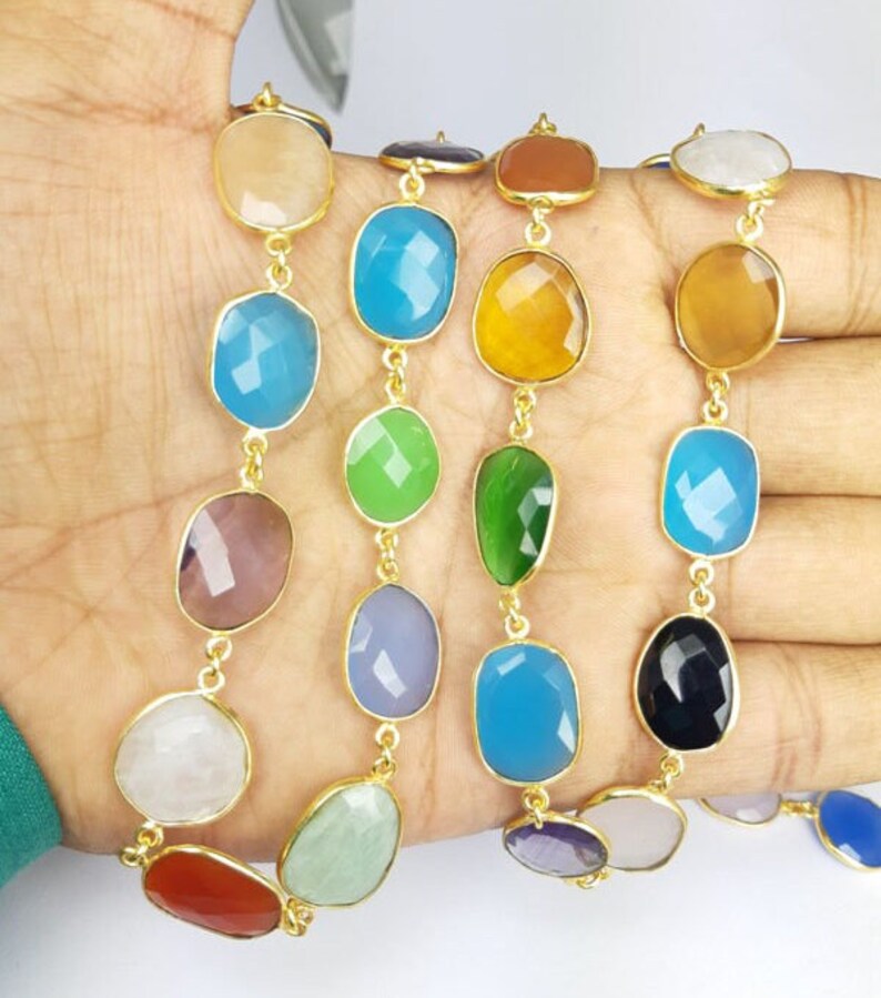 May include: A multi-colored gemstone necklace with gold-colored metal links. The necklace features a variety of faceted, oval-shaped gemstones in shades of blue, green, yellow, and purple. The gemstones are approximately 1 inch in length.