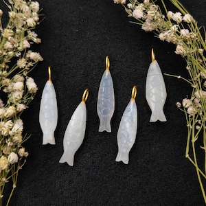 Puede incluir: Cinco colgantes de pez blanco translúcido con aros dorados para colgar. Los colgantes están tallados con detalles de escamas y dispuestos sobre una superficie negra, con flores blancas en el fondo. Estos colgantes son adecuados para la fabricación de joyas.
