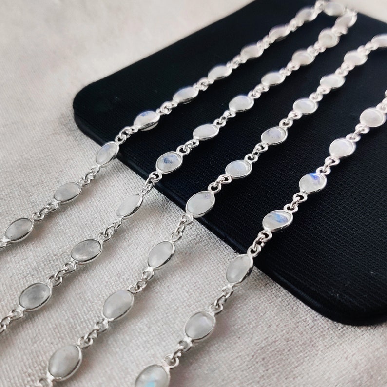 May include: Four silver chains with oval-shaped moonstone beads. The chains are laid out on a black surface.