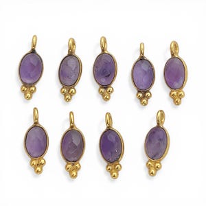 Può includere: Una collezione di nove pendenti color oro, ciascuno con una pietra di ametista ovale e sfaccettata. I pendenti hanno un piccolo anello nella parte superiore e un gruppo di tre piccole perle d'oro nella parte inferiore. Le ametiste sono di colore viola intenso.