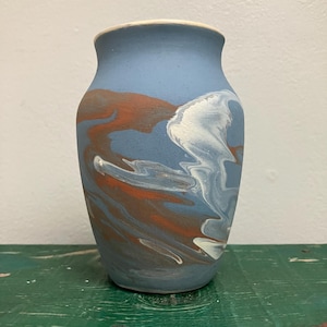 May include: A matte light blue vase featuring a marbled pattern of swirling white and terracotta orange paint across its surface. The vase has a rounded body, a slightly flared neck, and a smooth, unglazed rim at the top opening.