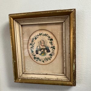 May include: An antique framed print featuring a woman in a white bonnet and blue and purple clothing, surrounded by a floral wreath. The frame is gold with a distressed finish, adding to the vintage aesthetic.