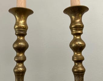 Könnte beinhalten: Ein Paar hohe Messingkerzenständer mit komplizierten geätzten Mustern entlang der Schäfte und ausgestellten Tropfschalen. Jeder Kerzenständer hält eine lange, schlanke, helle Stabkerze und zeigt ein metallisches Gold-Finish.
