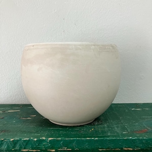 May include: A light beige, rounded ceramic bowl with a wide opening. The bowl has a smooth, matte finish and sits on a dark green, weathered wooden surface. The background is a plain white wall.