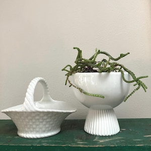May include: Two white ceramic decorative pieces. A basket with a handle and a textured basket weave design. A pedestal planter with a round bowl and a fluted base, holding a green plant with trailing stems.