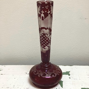 May include: A ruby red glass vase with etched designs. The vase has a bulbous base with a slender, elongated neck. The etched patterns include floral and architectural motifs. The vase is approximately 10 inches tall.