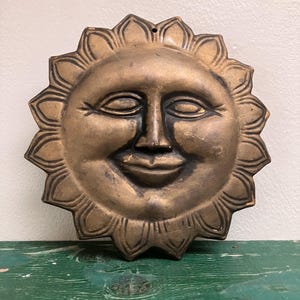 May include: A bronze-colored sun face wall hanging with a smiling expression. The sun has a detailed face with closed eyes and a smiling mouth. The sun's rays are stylized with a petal-like design. The wall hanging is made of metal.