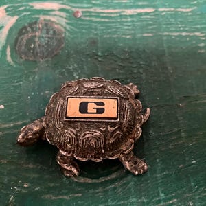 May include: A metallic silver-toned turtle figurine featuring a textured shell with a rectangular inset on the back. The inset is light tan with a bold, dark navy blue capital letter G centered on its surface. The turtle has a detailed head and legs.