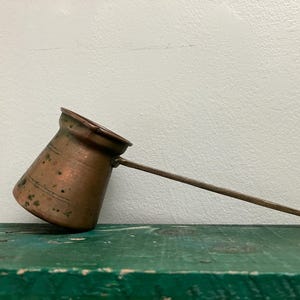 May include: A copper coffee pot with a long, slender metal handle. The pot has a tapered, cylindrical body with a wide, flared rim and a distinct pouring spout. The exterior surface shows a weathered, oxidized patina with dark spots.