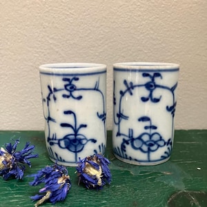 May include: Two white ceramic cups with blue floral patterns. The cups are cylindrical and have a blue rim. Dried blue flowers are scattered around the cups on a green surface. The cups have a classic, vintage aesthetic.