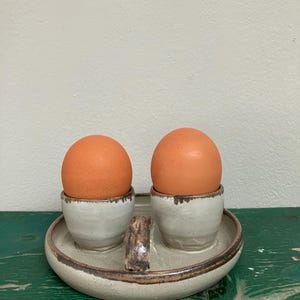 May include: A ceramic egg holder with two eggs. The eggs are brown and sit in small, white and gray cups. The cups are on a round, gray tray with a handle. The tray has a brown rim.