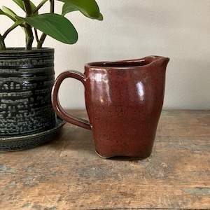 May include: A red speckled ceramic pitcher with a handle. The pitcher has a rounded body and a slightly flared lip.