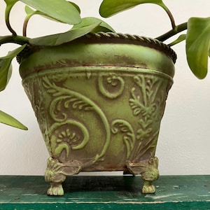 May include: A green metal planter with ornate embossed floral designs and a rope-like trim. The planter sits on three decorative feet and holds a leafy green plant. The planter has a distressed finish, giving it a vintage look.