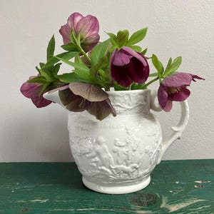 May include: A white ceramic mug with a black handle and the text "But First, Coffee" in a bold, black font. The mug is set against a blurred background, suggesting a cozy setting.