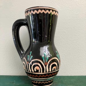 May include: A glossy black ceramic pitcher featuring a hand-painted decorative pattern. The design includes cream-colored zig-zag lines at the rim, repeating arched motifs with orange dots, and teal leaf-like accents around the rounded body.