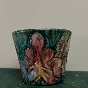 May include: A small, handcrafted ceramic pot with a textured surface. The pot features a teal green exterior with a central design of pink and red floral shapes. Additional colors include yellow, blue, and brown.