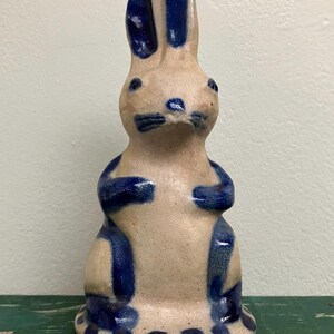 May include: A ceramic rabbit figurine featuring a light tan, unglazed clay body with hand-painted cobalt blue accents on the ears, eyes, nose, whiskers, paws, legs, and a series of circular dots around the base of the upright, seated animal.