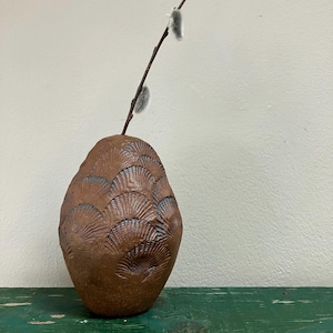 May include: A brown ceramic vase with a textured, fan-like pattern. A slender branch with two fluffy, gray catkins extends from the top. The vase is on a green surface against a white wall.