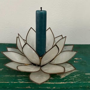 May include: A white lotus flower candle holder with a teal candle. The candle holder has a brass-colored trim and is sitting on a green surface. The candle is a solid teal color.