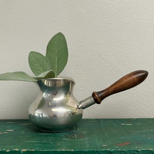 May include: A polished silver-toned metal coffee pot with a rounded body and a long, dark brown wooden handle. Several broad, fuzzy, light green sage leaves are tucked inside the opening of the pot, extending upwards from the metallic vessel.