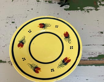 Puede incluir: Un plato de cerámica amarillo con borde negro y diseño floral. El plato presenta un círculo negro central y está decorado con motivos florales rojos, amarillos y verdes. Una figura de pájaro de metal oscuro está colocada encima del plato.