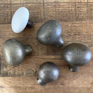 May include: A collection of five antique cabinet knobs. One is white and oval-shaped, while the other four are metallic gray with a rounded, mushroom-like design. The knobs are arranged on a wooden surface, showcasing their vintage aesthetic.