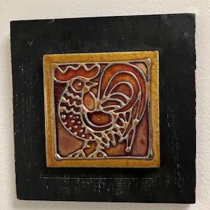 May include: A square, decorative tile featuring a rooster design in shades of orange, red, and brown, outlined in silver. The tile is set within a gold border and mounted on a black wooden base.