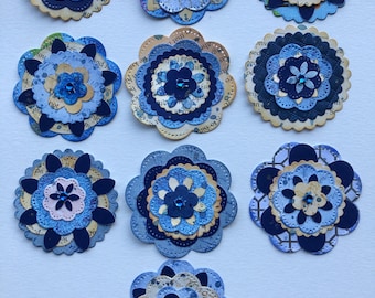 May include: Nine handmade paper flowers in shades of blue, with layered petals and decorative edges. Each flower features a central embellishment, some with a small gem. The flowers are arranged on a white surface.