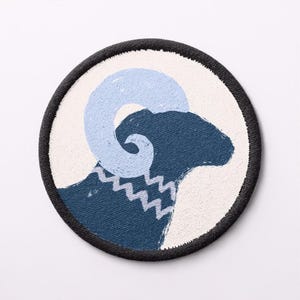 May include: A circular patch with a navy blue ram's head design and a light blue horn. The ram has a white zig-zag pattern on its neck, set against a white background, with a black border.