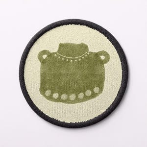May include: A circular patch with an olive green pot design, complete with handles and dotted embellishments, on a cream backdrop. The patch is framed by a black stitched border, a decorative element.