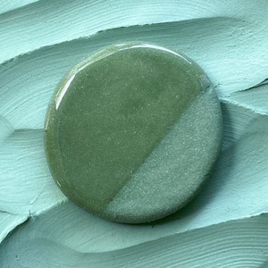 May include: A round ceramic tile with a green glaze on one half and a white glaze on the other half. The tile has a slightly textured surface.