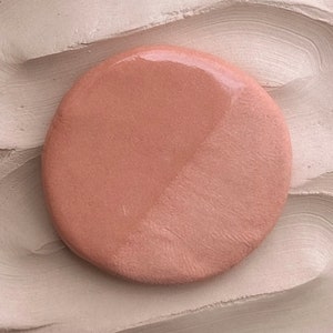 May include: A round, smooth, terracotta-colored clay disc with a slightly glossy finish. The disc is a light peach color on one side and a darker peach color on the other side.